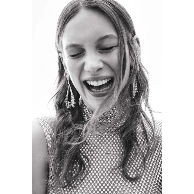 Close-up black and white portrait of a smiling young woman with long wavy hair, wearing a metallic top and dangling earrings.