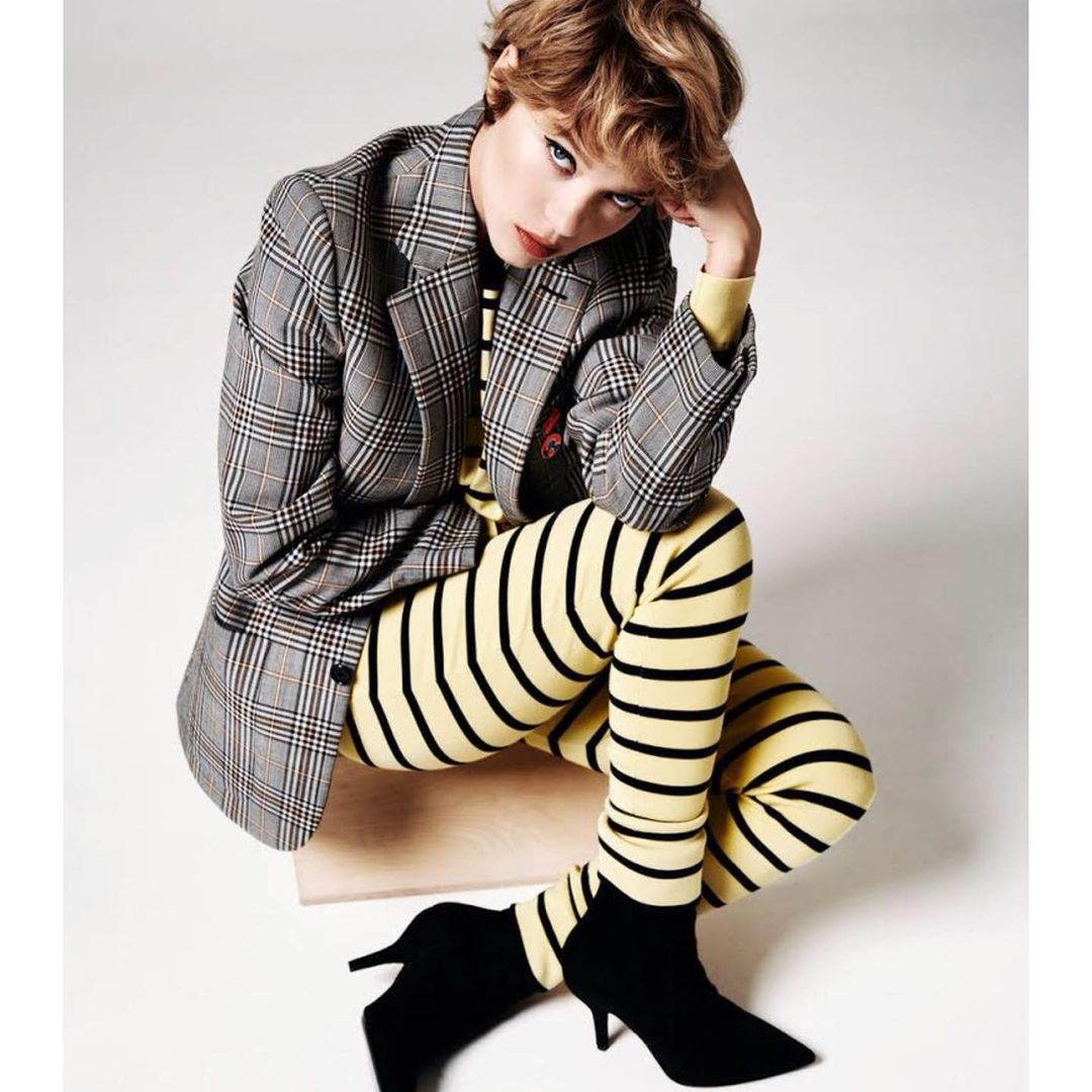 A fashion portrait of a young woman with short hair, wearing a blazer, striped leggings, and heels, seated in a photo studio setting.