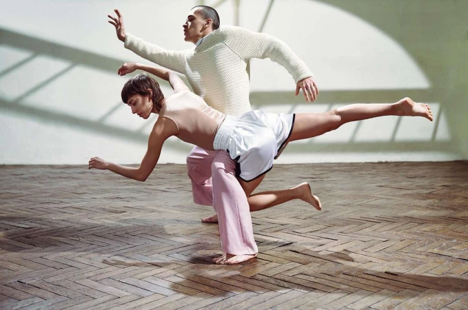 Two dancers in motion on a parquet wooden floor with dramatic window shadows on the back wall
