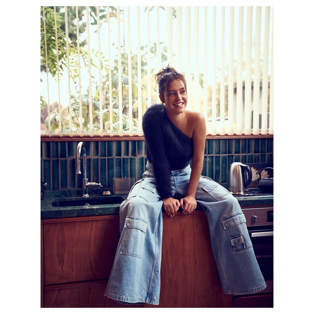 Young woman with a joyful smile sitting on a wooden counter in a kitchen setting, wearing loose jeans and a one-shoulder knit top.