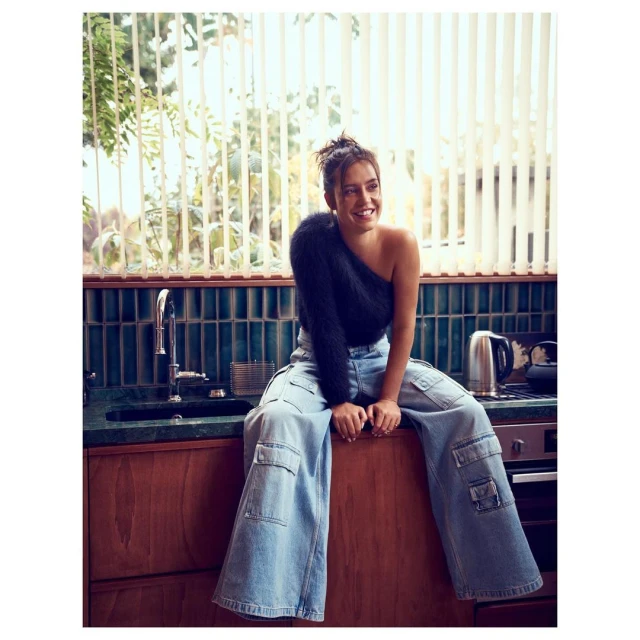 Young woman with a joyful smile sitting on a wooden counter in a kitchen setting, wearing loose jeans and a one-shoulder knit top.