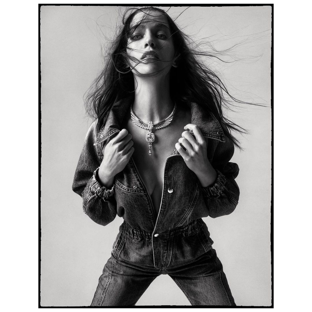 Monochrome portrait of a woman with windswept hair, wearing a denim jumpsuit and statement necklace against a neutral backdrop.
