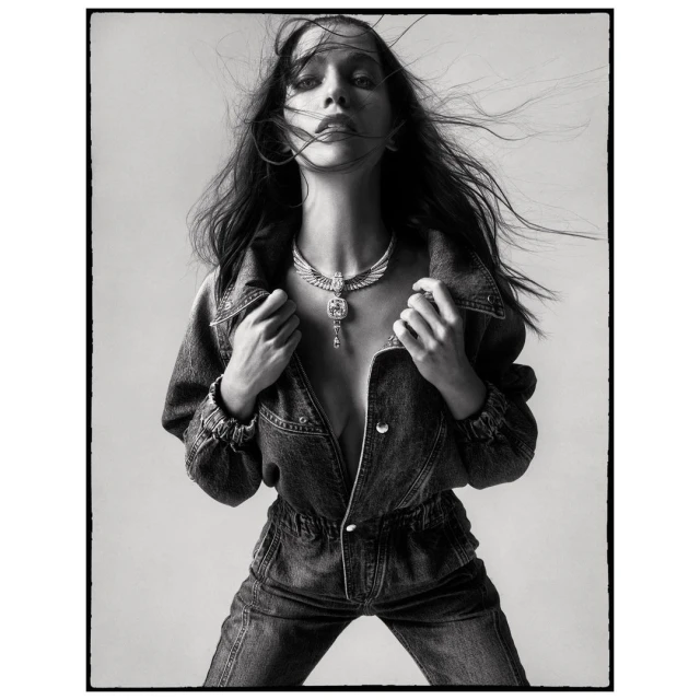 Monochrome portrait of a woman with windswept hair, wearing a denim jumpsuit and statement necklace against a neutral backdrop.