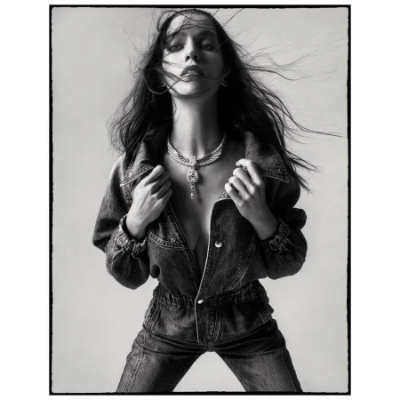 Monochrome portrait of a woman with windswept hair, wearing a denim jumpsuit and statement necklace against a neutral backdrop.