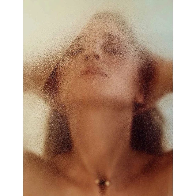 Portrait of a woman seen through a steamed-up glass shower door, with her hands behind her head, showing a necklace.