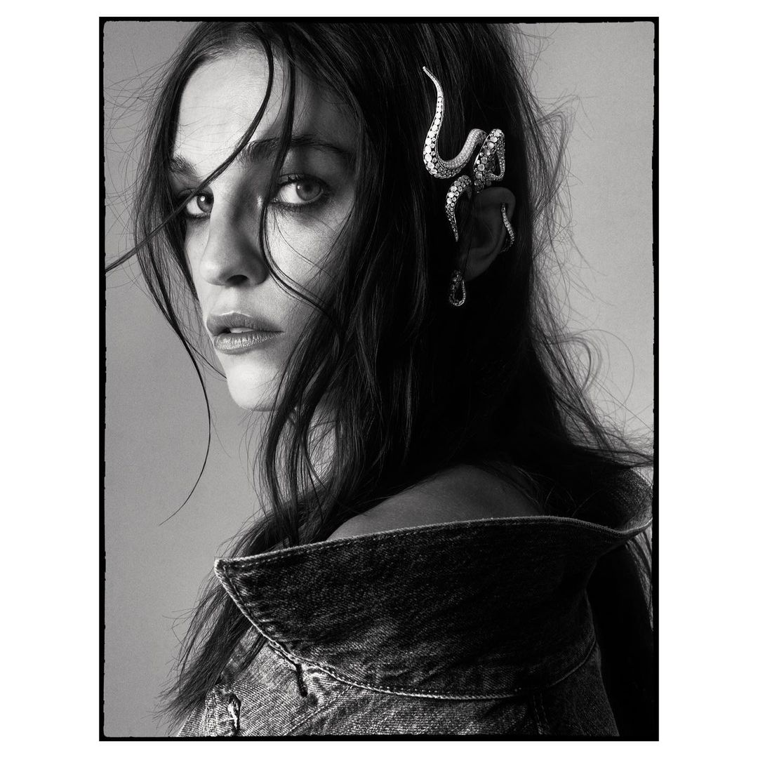 Black and white portrait of a female model wearing a denim jacket and unique snake earring, gazing intently at the camera.