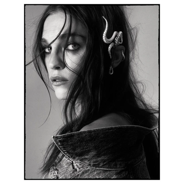 Black and white portrait of a female model wearing a denim jacket and unique snake earring, gazing intently at the camera.