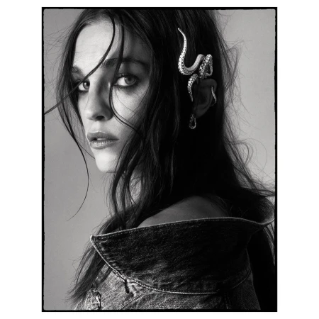 Black and white portrait of a female model wearing a denim jacket and unique snake earring, gazing intently at the camera.