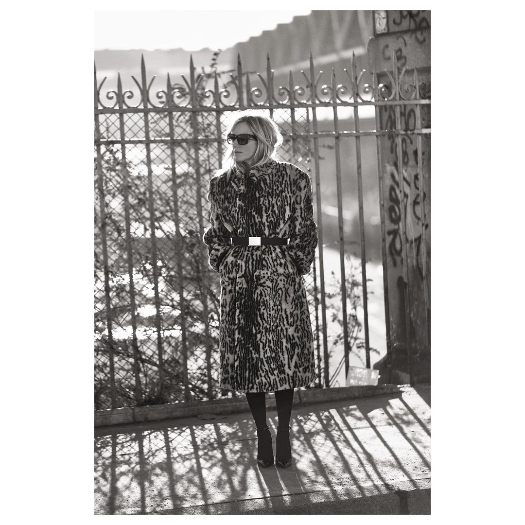 A solitary adult female stands in an urban setting, modeling a leopard print coat, sunglasses, and boots against a bridge and metal fence.