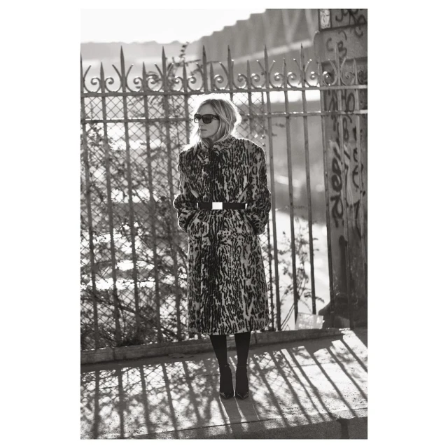 A solitary adult female stands in an urban setting, modeling a leopard print coat, sunglasses, and boots against a bridge and metal fence.