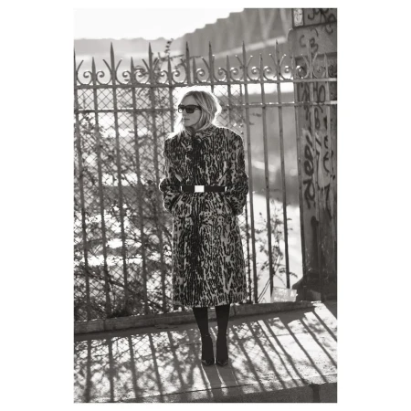 A solitary adult female stands in an urban setting, modeling a leopard print coat, sunglasses, and boots against a bridge and metal fence.