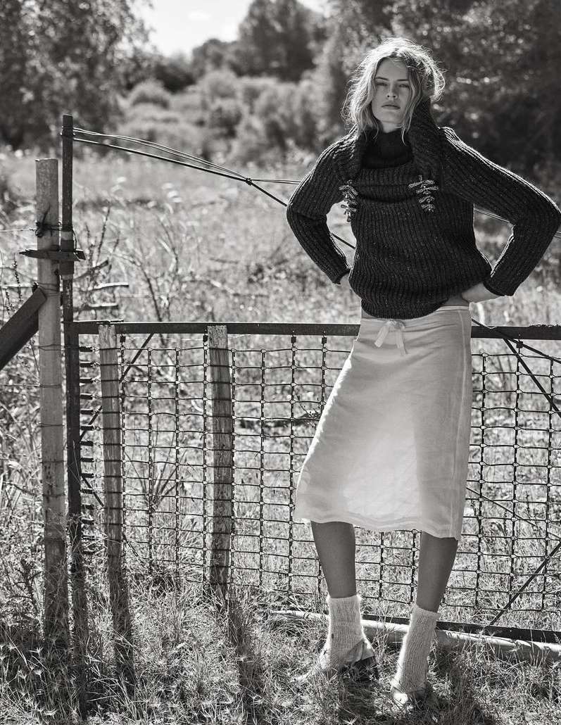 Monochrome portrait of a slender blonde woman posing near a weathered metal fence in a rural setting, wearing a knit sweater and skirt.
