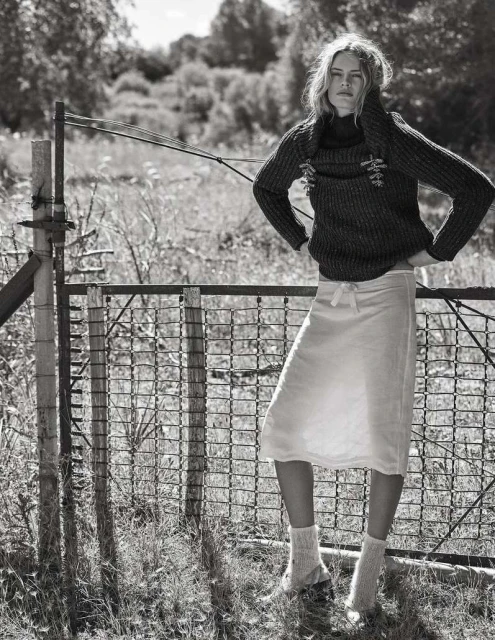 Monochrome portrait of a slender blonde woman posing near a weathered metal fence in a rural setting, wearing a knit sweater and skirt.