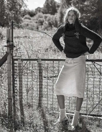 Monochrome portrait of a slender blonde woman posing near a weathered metal fence in a rural setting, wearing a knit sweater and skirt.