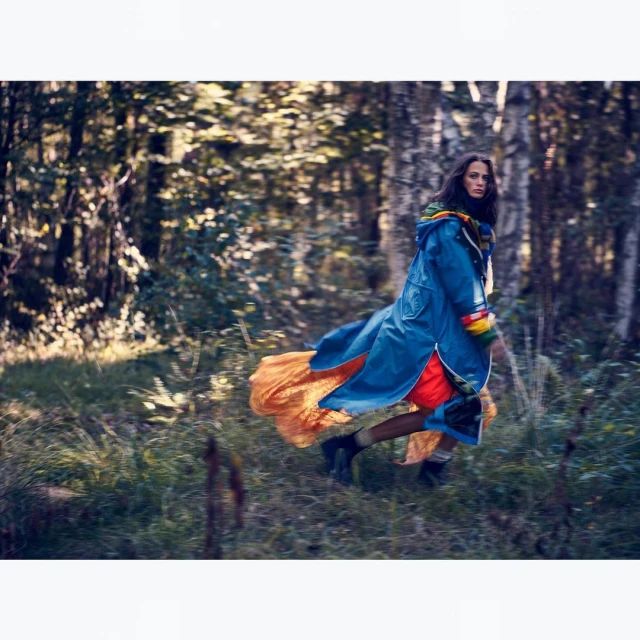 A young adult woman runs through a sunlit forest clearing wearing a blue raincoat and orange dress, captured in a dynamic editorial style.