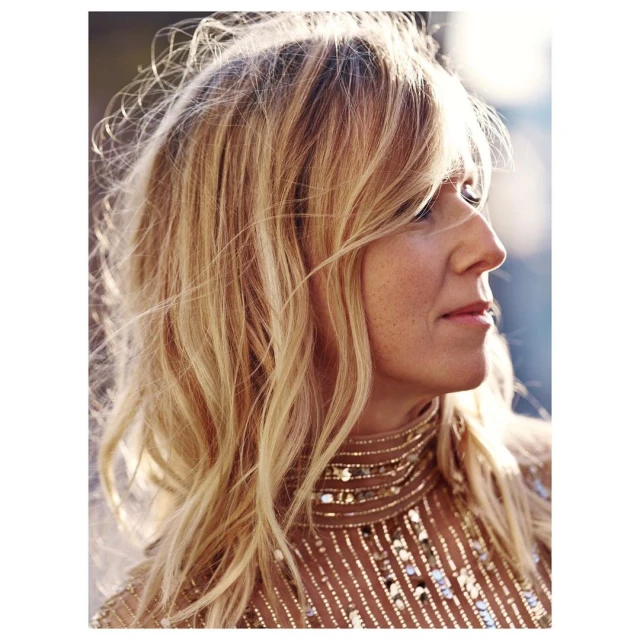 Blonde woman in glittering top gazes to the right in a close-up shot capturing her wavy hair and the shimmering detail of her high-necked garment.