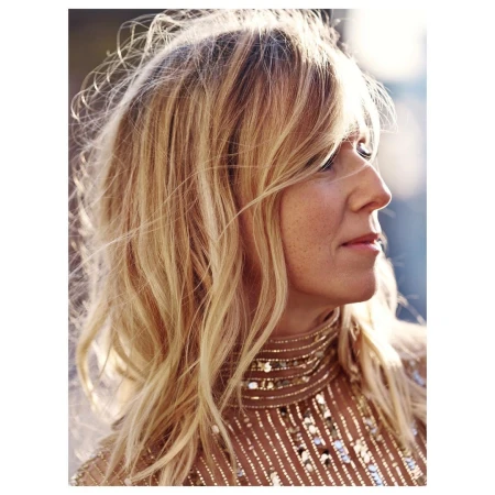 Blonde woman in glittering top gazes to the right in a close-up shot capturing her wavy hair and the shimmering detail of her high-necked garment.