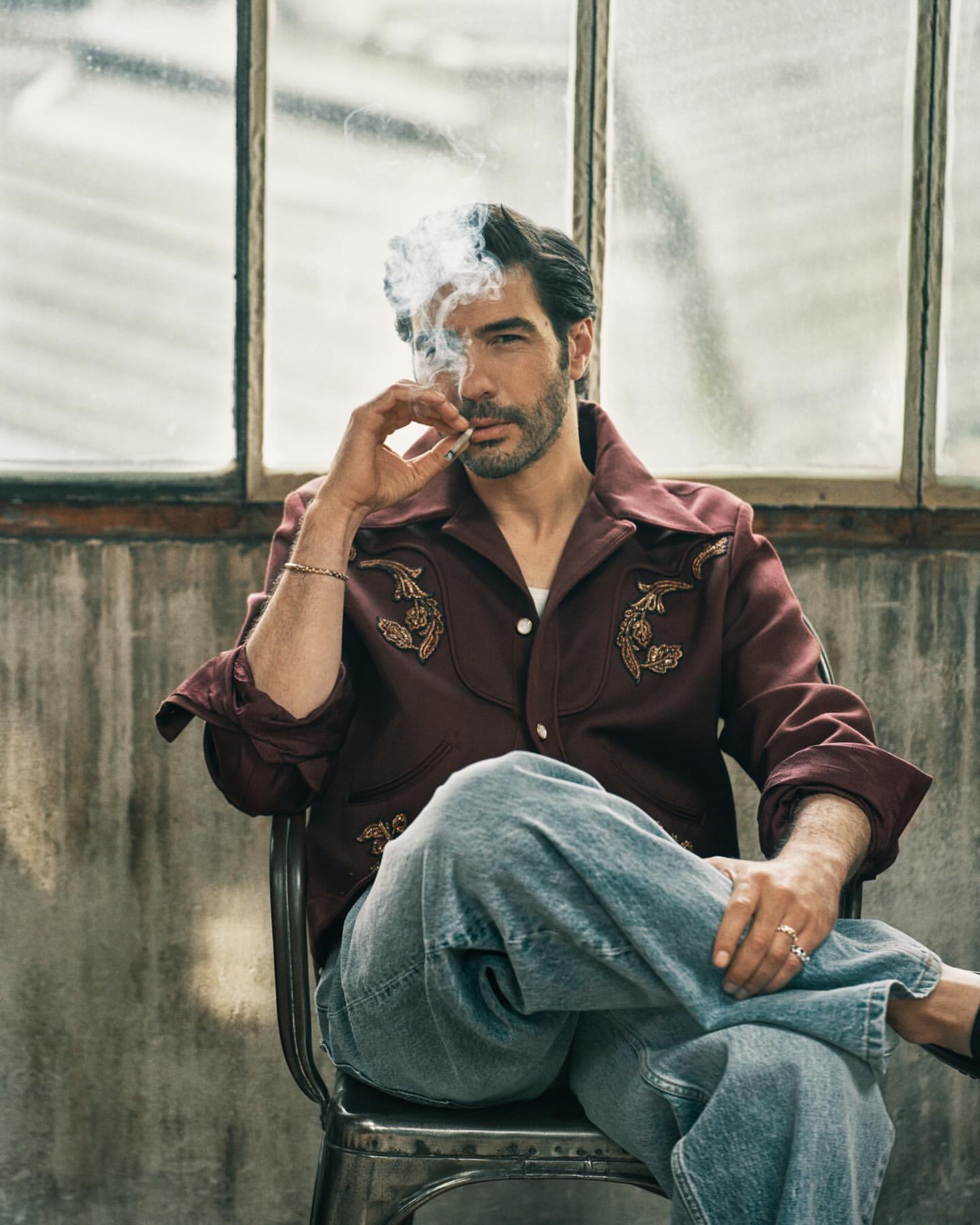 Portrait of a man with dark hair and a beard wearing a western style shirt and jeans sitting on a metal chair and smoking.