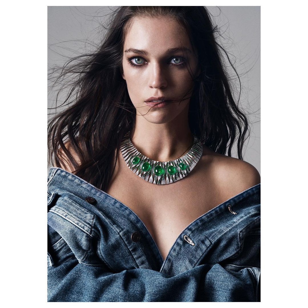 A striking portrait of a woman with dark hair wearing a statement necklace and a denim jacket against a soft grey backdrop.