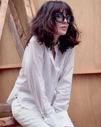 Adult woman with dark hair and sunglasses wearing a white shirt and pants, posing in front of a wooden background indoors.