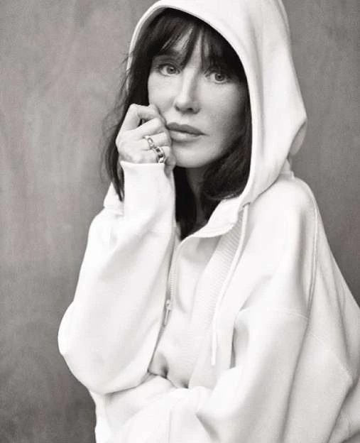 Monochrome portrait of a woman with dark hair wearing a hooded sweatshirt, hand near her face, set against a gray background.