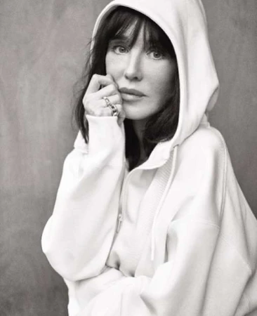 Monochrome portrait of a woman with dark hair wearing a hooded sweatshirt, hand near her face, set against a gray background.