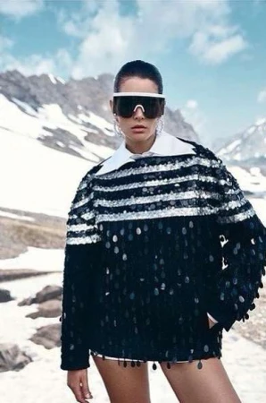 Brunette woman in oversized sequin sweater and large sunglasses in mountain environment under a bright blue sky with clouds.