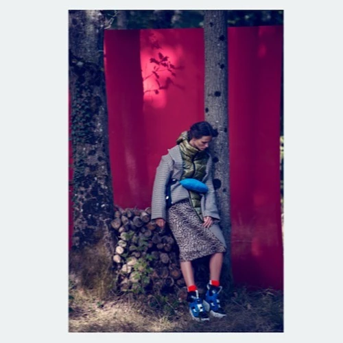 A woman in a coat leans against a tree, wearing a blue fanny pack and athletic shoes, in a forest with red background panels.