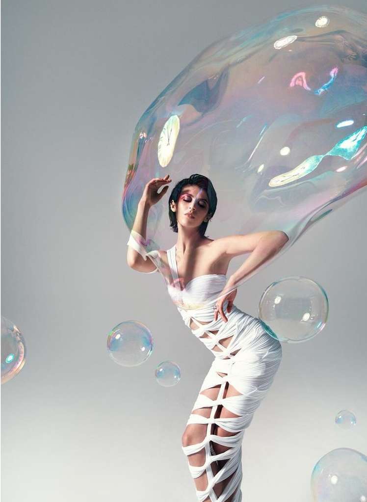 Woman in a one-shoulder dress posing with a large iridescent soap bubble and surrounding smaller bubbles in a studio environment.