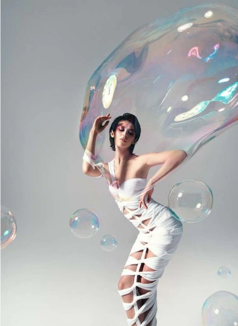 Woman in a one-shoulder dress posing with a large iridescent soap bubble and surrounding smaller bubbles in a studio environment.