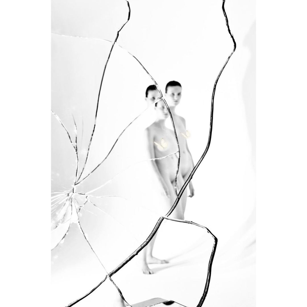 Two slender, nude figures standing behind a shattered mirror, set against a stark white backdrop in a bright photo studio setting.