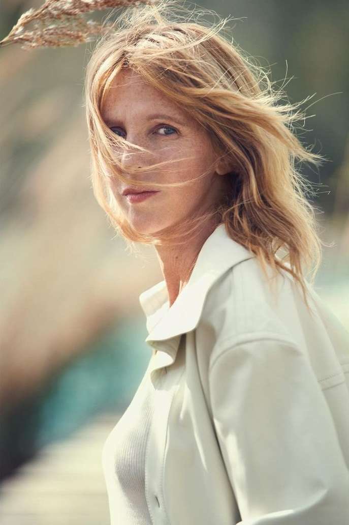 Portrait of a blonde woman with windswept hair in a light-colored jacket, gazing at the camera with a neutral expression in an outdoor setting.