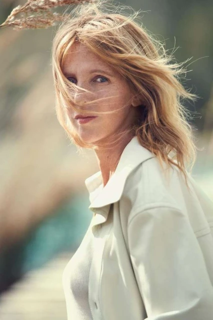 Portrait of a blonde woman with windswept hair in a light-colored jacket, gazing at the camera with a neutral expression in an outdoor setting.