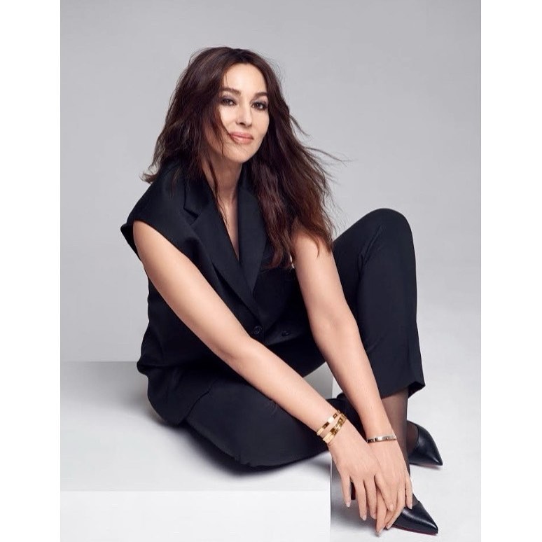 Monica Bellucci poses in black suit and heels against a gray background, accessorized with gold bracelets, with soft studio lighting.