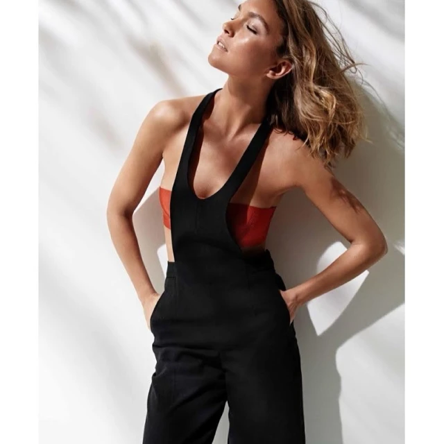 Woman in black overalls and an orange top, lit by the sun, casting a shadow on the white wall behind her. Casual fashion.