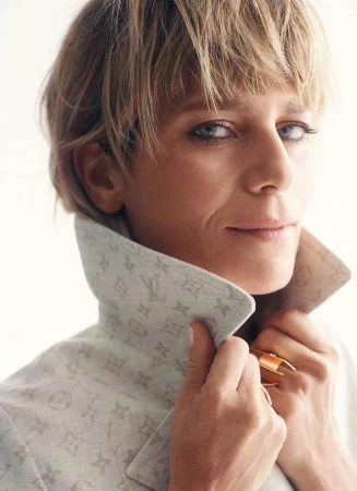 Close-up portrait of a woman with short blonde hair wearing a patterned high-collar coat against a bright white background.