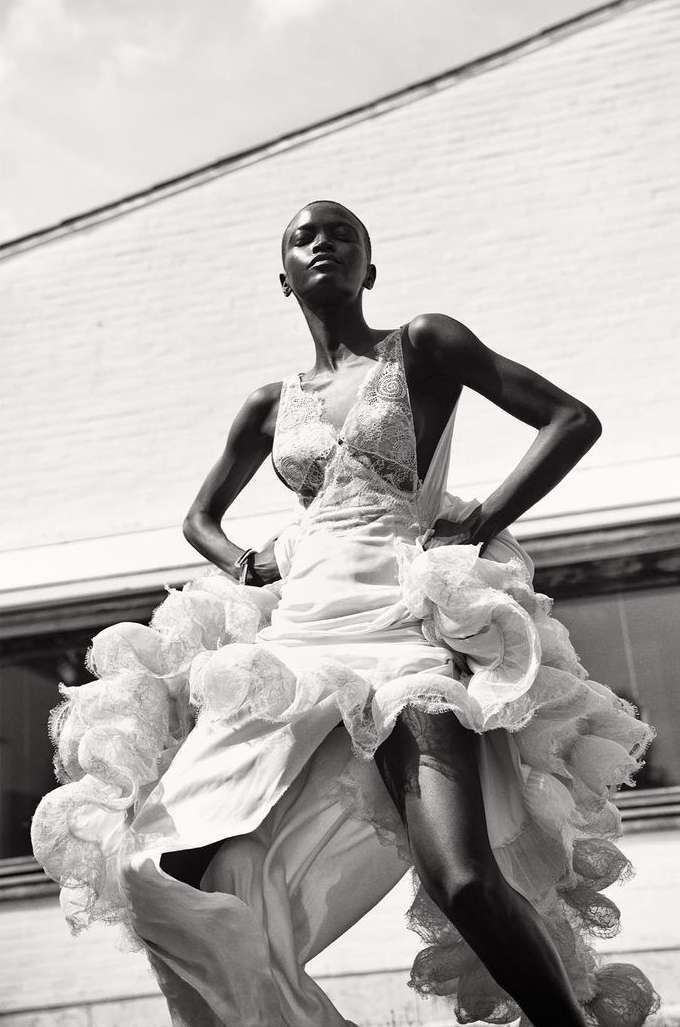 Fashion shot of a slender Black woman in an ornate white gown, confidently posing outdoors against a neutral building under bright sky.