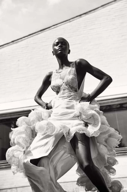 Fashion shot of a slender Black woman in an ornate white gown, confidently posing outdoors against a neutral building under bright sky.