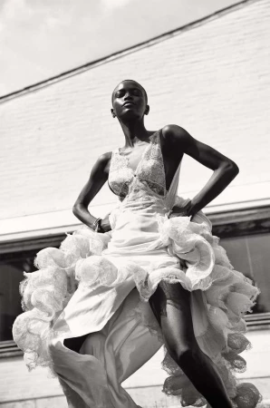 Fashion shot of a slender Black woman in an ornate white gown, confidently posing outdoors against a neutral building under bright sky.