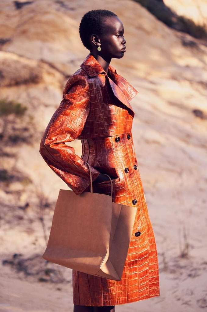 A fashion editorial shot of a Black woman in an orange coat standing outside holding a shopping bag in a tan environment.