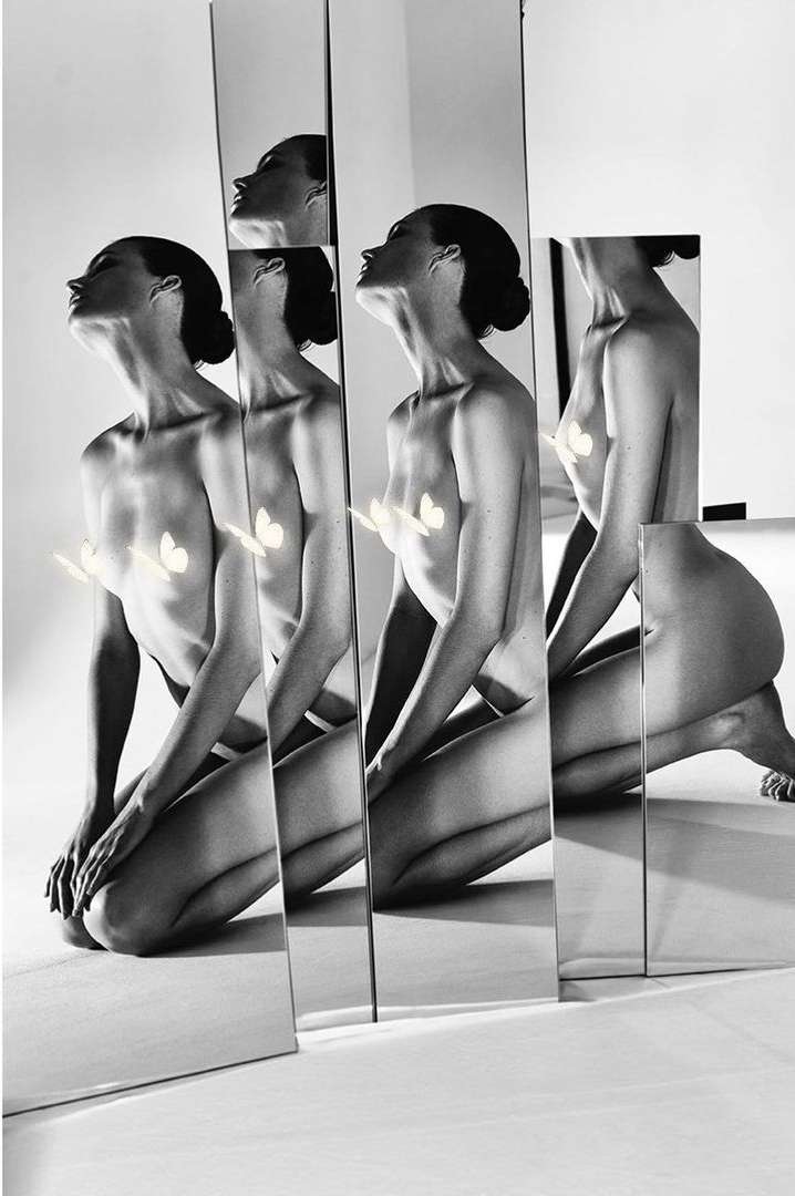 Artistic black and white image featuring a nude woman kneeling behind several vertical mirrors reflecting her form in a photo studio.