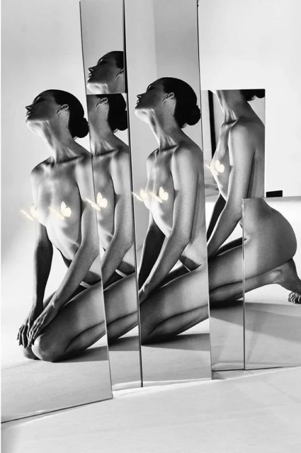 Artistic black and white image featuring a nude woman kneeling behind several vertical mirrors reflecting her form in a photo studio.