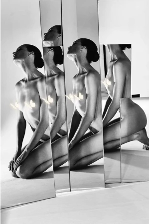 Artistic black and white image featuring a nude woman kneeling behind several vertical mirrors reflecting her form in a photo studio.