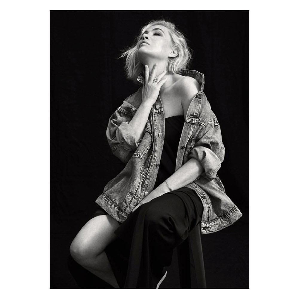 Monochrome portrait of woman with blonde hair and denim jacket, seated in studio with black backdrop, looking up.