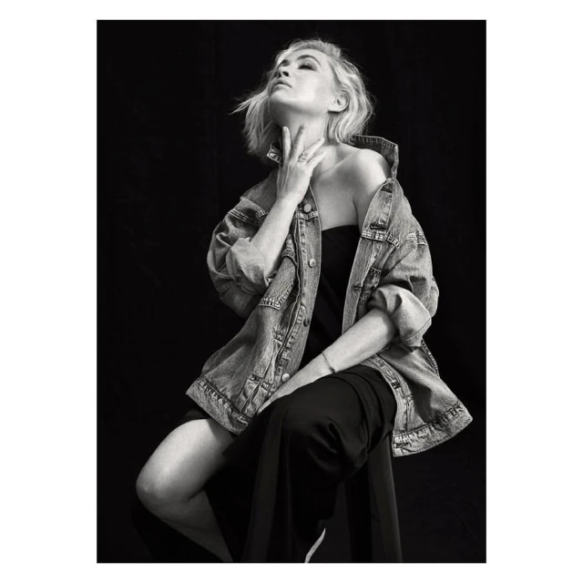 Monochrome portrait of woman with blonde hair and denim jacket, seated in studio with black backdrop, looking up.
