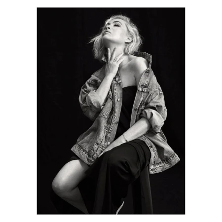 Monochrome portrait of woman with blonde hair and denim jacket, seated in studio with black backdrop, looking up.