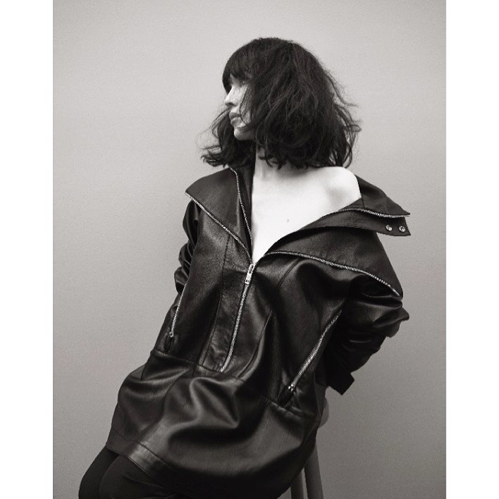 Monochrome portrait of a woman with dark hair wearing an oversized leather jacket against a plain background.