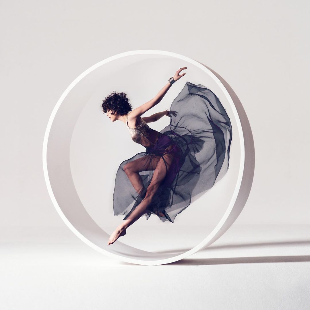 Adult female in sheer dress leaps through a large white ring in a studio with soft lighting and visible shadow, performing a dance pose.
