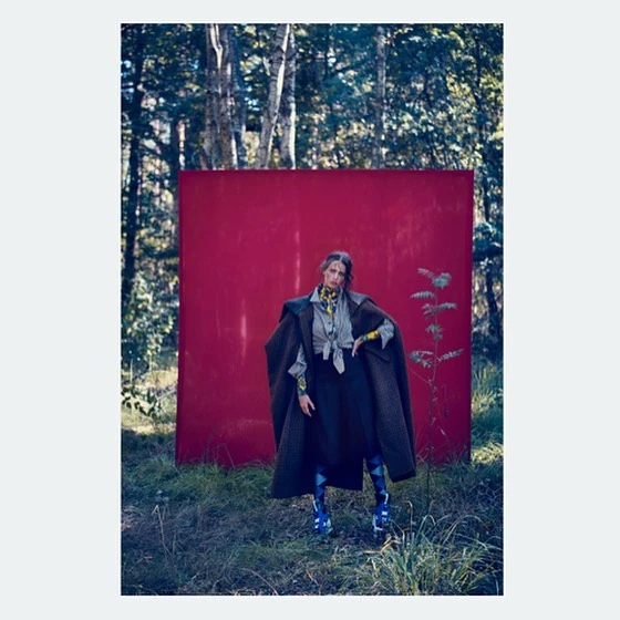 Fashionable adult female in a green coat poses in a forest with a large red background, showcasing nature-inspired editorial style.