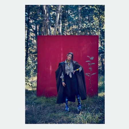 Fashionable adult female in a green coat poses in a forest with a large red background, showcasing nature-inspired editorial style.